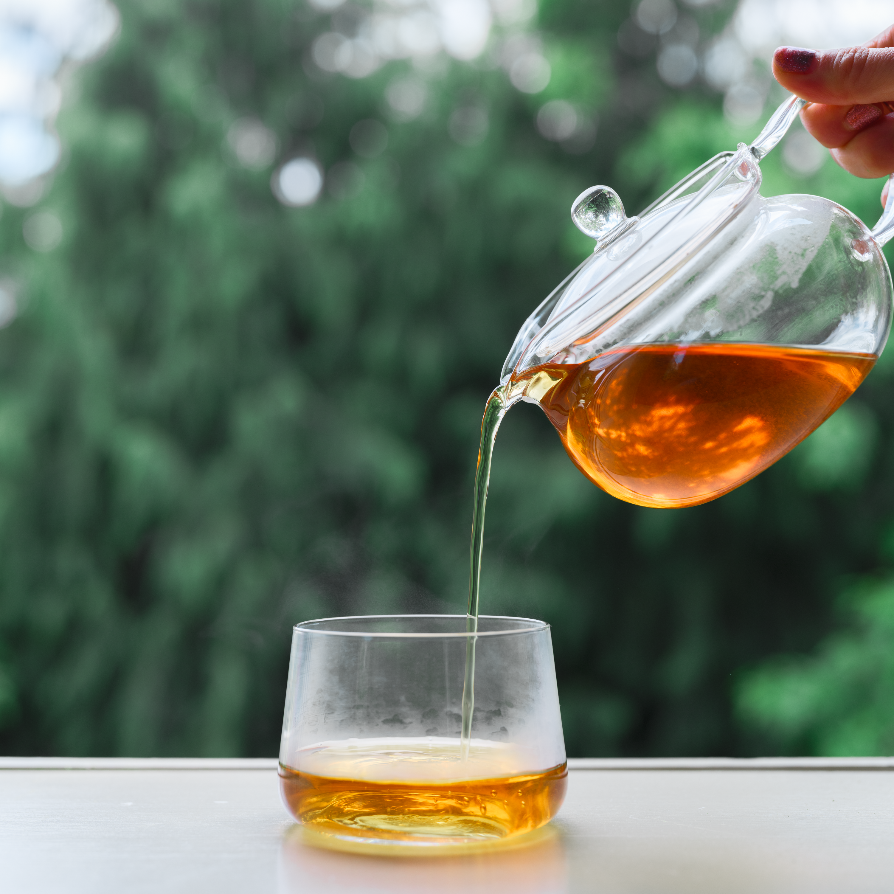 Glass teapot pouring tea into a glass with a blurred green background