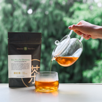 Person pouring Japanese tea from a glass teapot into a glass with a Mizuba Tea Co. Shincha package in the background.