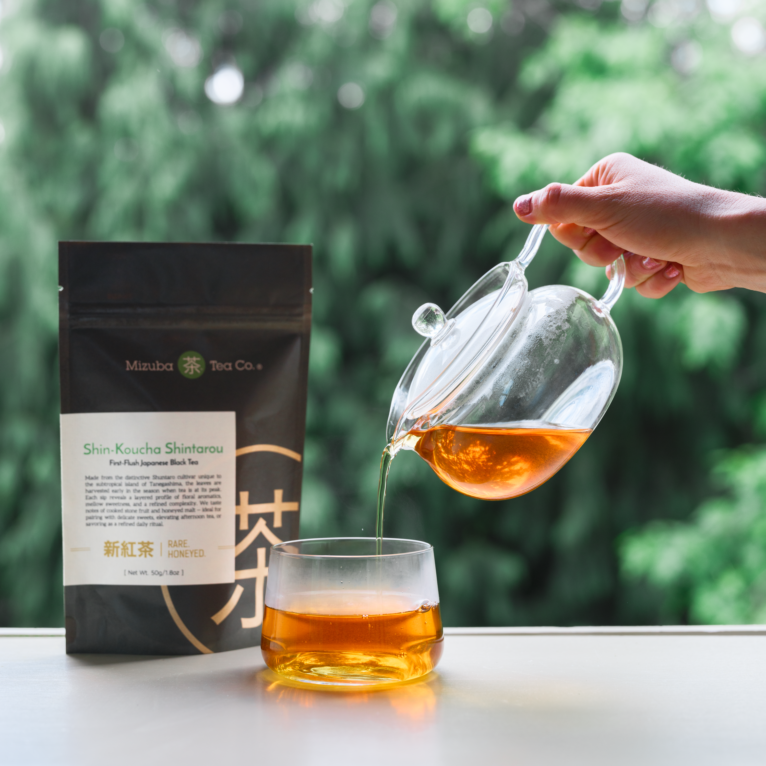 Person pouring Japanese tea from a glass teapot into a glass with a Mizuba Tea Co. Shincha package in the background.