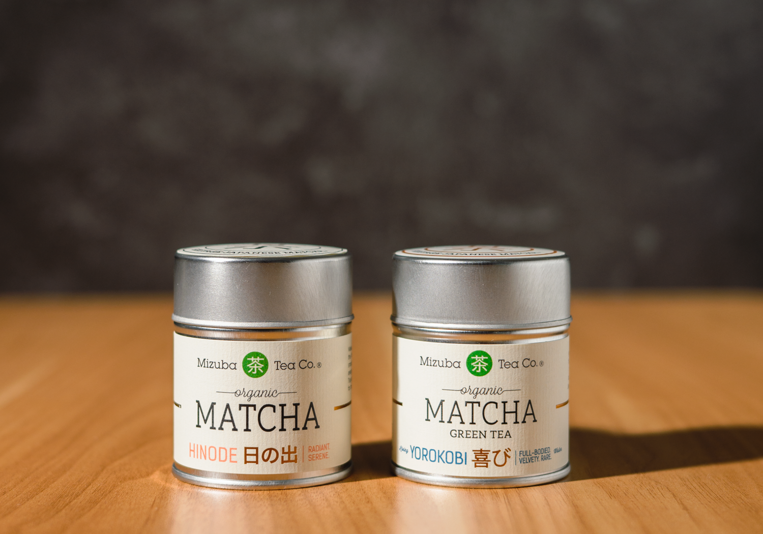 Two tins (Hinode and Yorokobi) of Mizuba Tea Co. Matcha green tea on a wooden surface with a dark background.