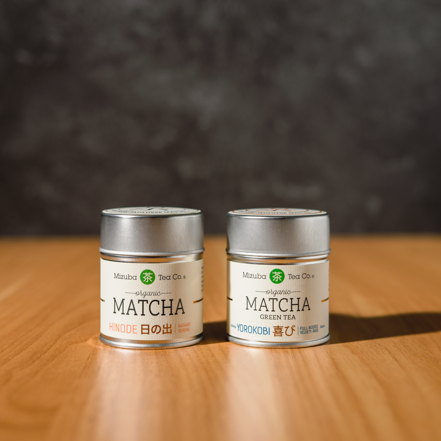 Two tins (Hinode and Yorokobi) of Mizuba Tea Co. Matcha green tea on a wooden surface with a dark background.