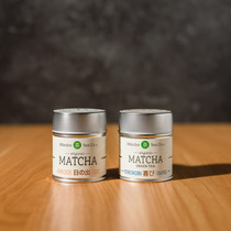 Two tins (Hinode and Yorokobi) of Mizuba Tea Co. Matcha green tea on a wooden surface with a dark background.