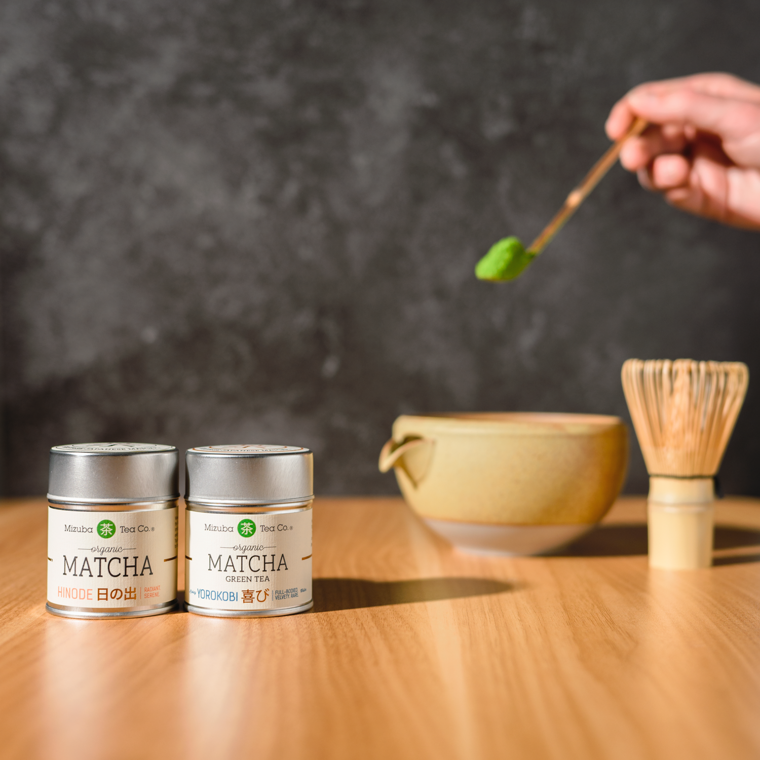 Two Mizuba Matcha green tea tins on a wooden table with a person preparing matcha.