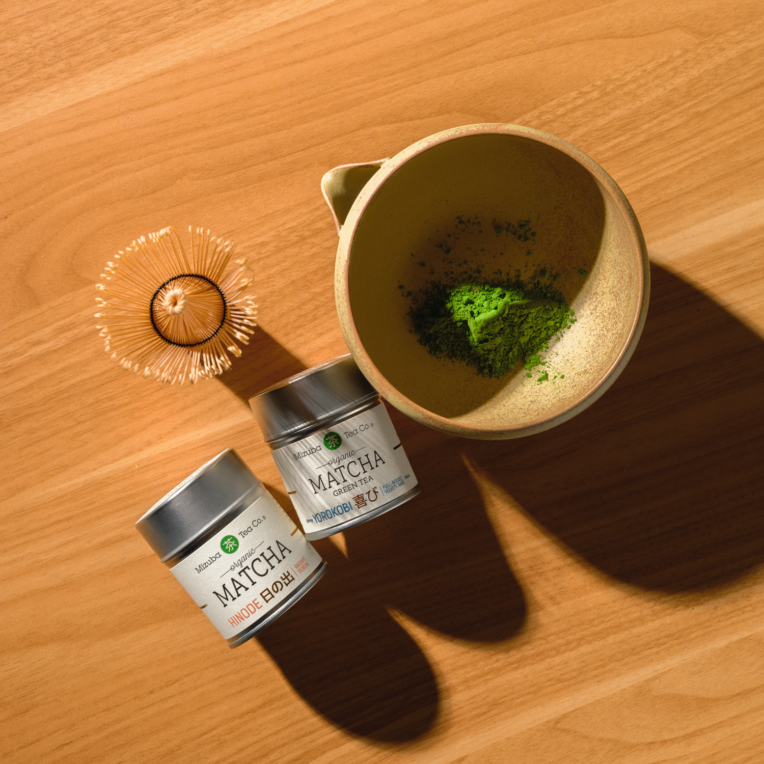 Mizuba Matcha powder in a bowl with two tins - Hinode and Yorokobi  – on a wooden surface.