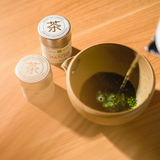Matcha being prepared in a yellow chawan tea bowl next to two tins of Mizuba Matcha tea on a wooden surface