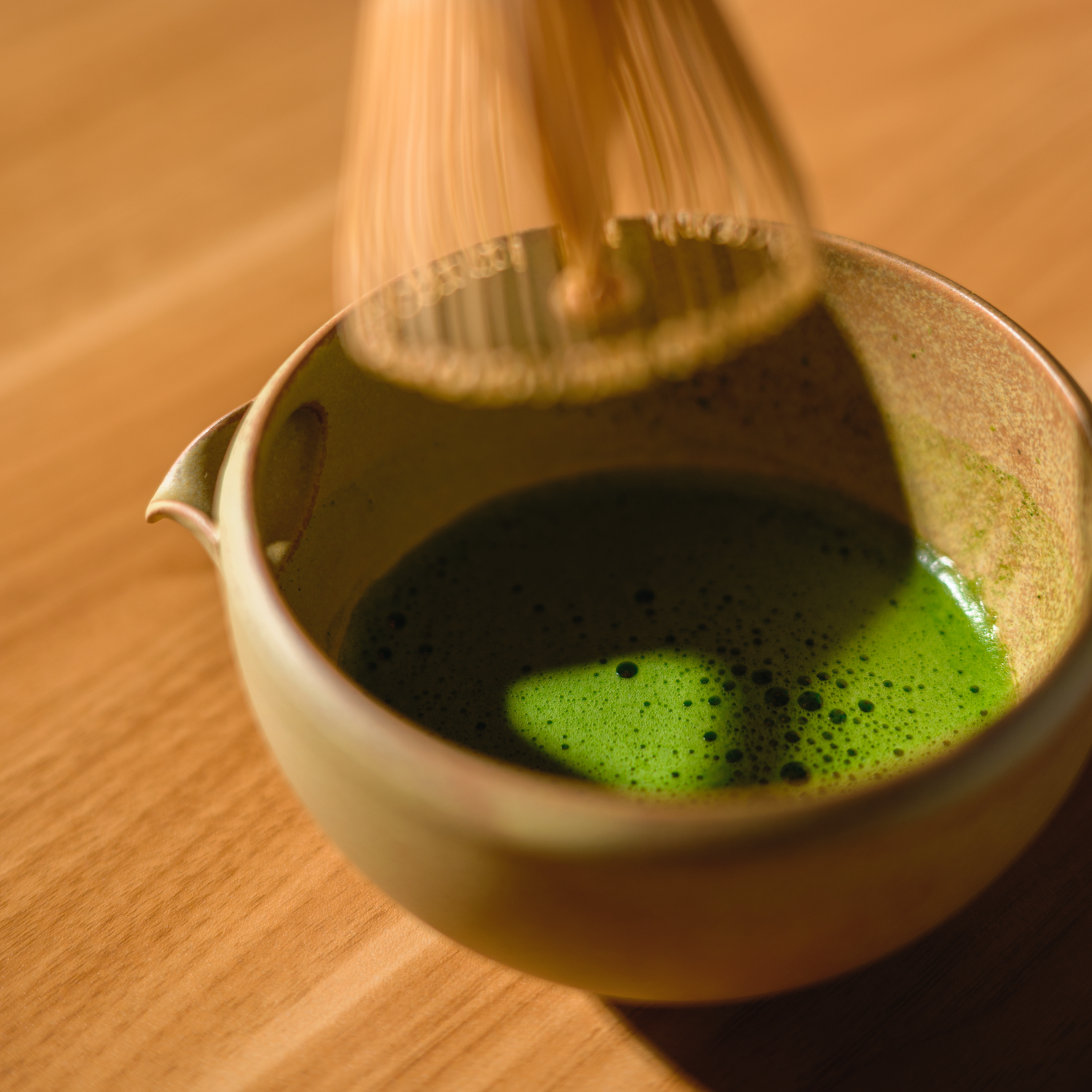 Mizuba matcha green tea is being whisked in a ceramic chawan tea bowl with a bamboo chasen whisk on a wooden surface.