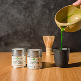 On the right, Mizuba Matcha green tea is poured from a chawan tea bowl into a black cup next to two Mizuba matcha tins on a wooden surface.