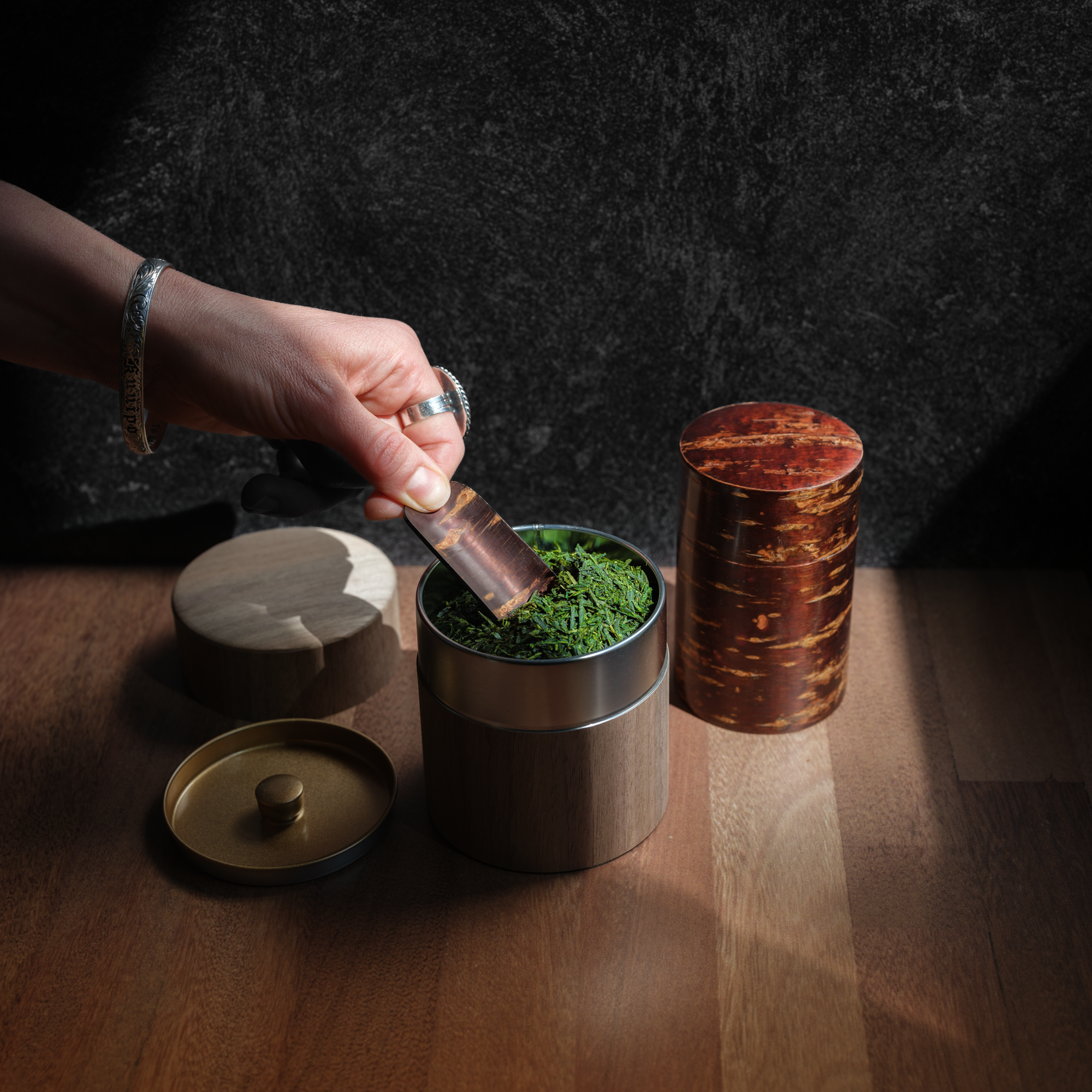 Person uses a cherry wood sakura chasaji tea scoop to lift sencha tea leaves from a walnut wrapped canister. 