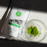 Mizuba Matcha powder in a clear glass chawan (tea bowl) with a package labeled 'Signature Matcha Yame' on a light background.
