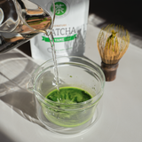Water intended to make matcha is poured into a glass chawan (tea bowl) filled with Yame First Flush matcha green tea with its packaging in the background. 