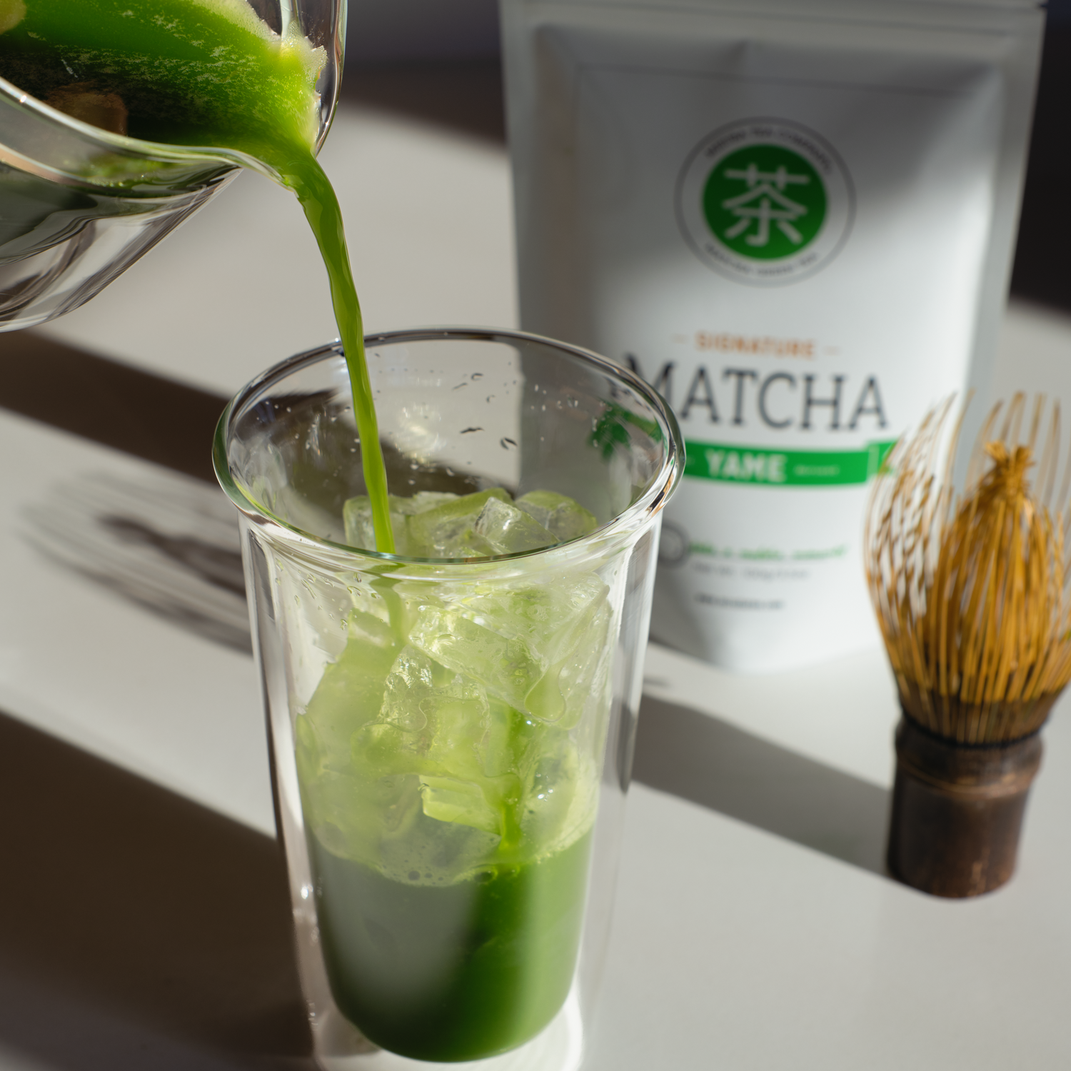 Matcha green tea being poured into a glass with Mizuba Yame First Flush matcha 100g pouch in the background.