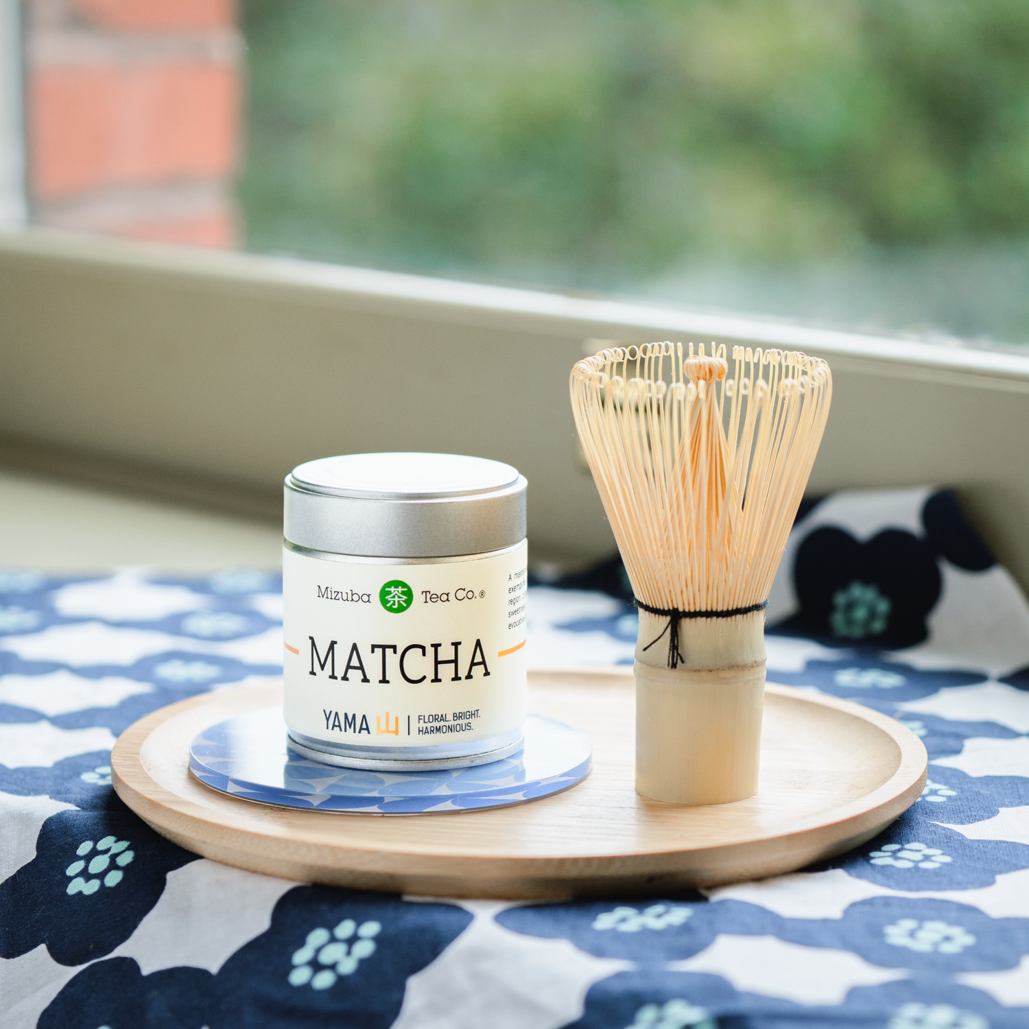 Mizuba Yama Matcha tea canister and chasen whisk on a wooden tray with a blue floral tablecloth.