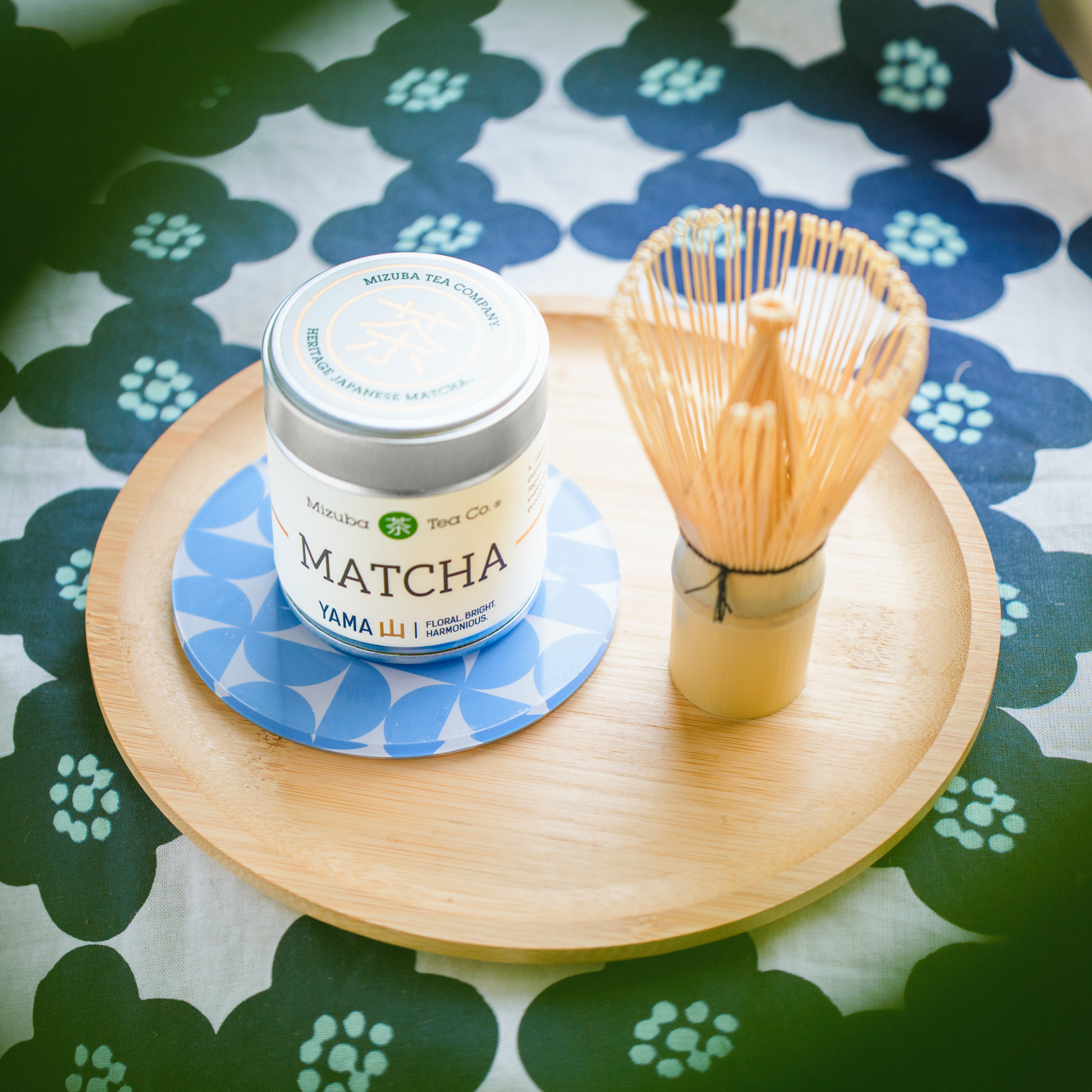 Mizuba Yama Matcha powder container with bamboo whisk on a wooden tray over a floral tablecloth.